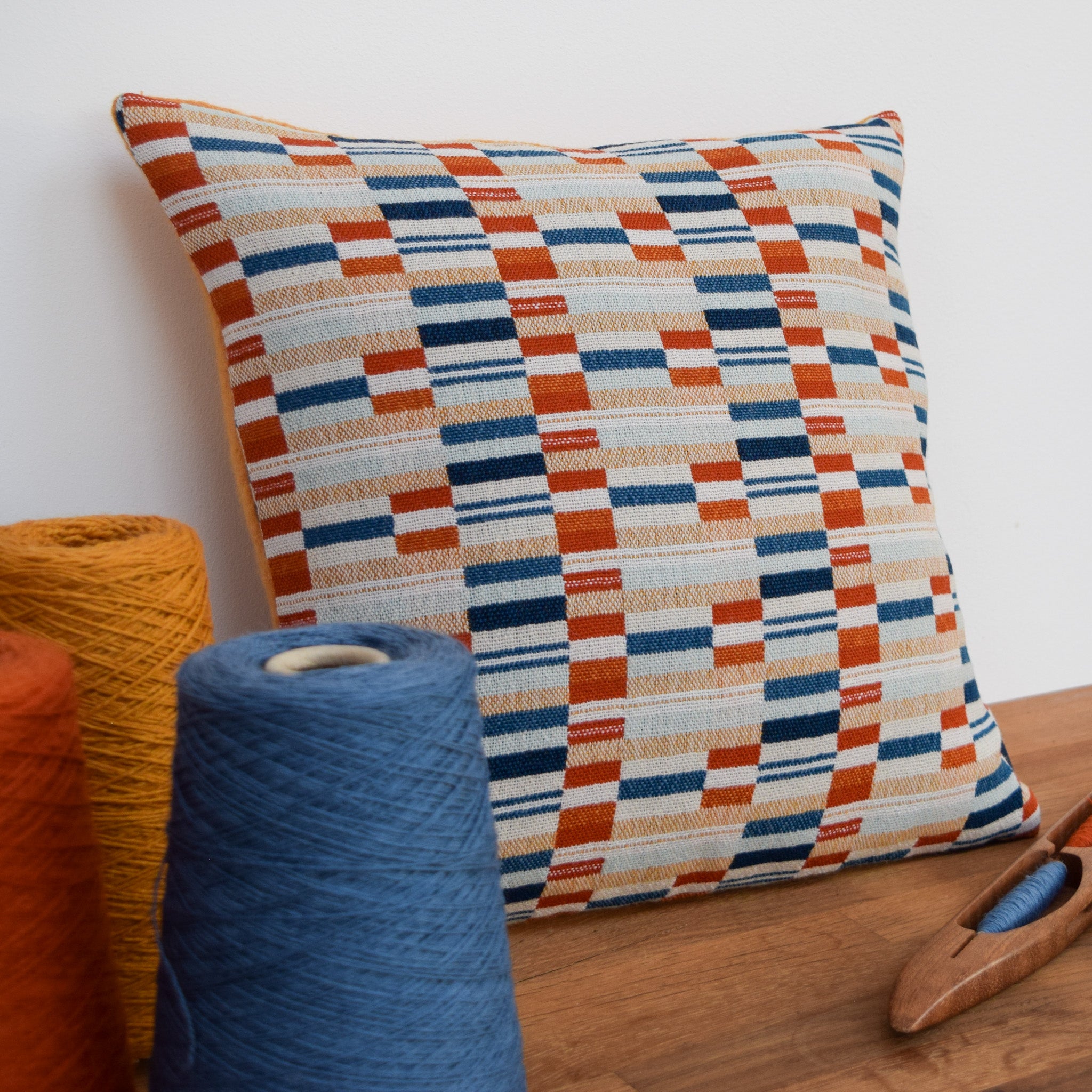 Photo showing a cushion with  a mixture of horizontal rectangular blocks of orange, yellow and blues to create the design. There are also three cones of yarn showing the burnt orange, mustard and blue yarns which have been used to create the handwoven cloth.