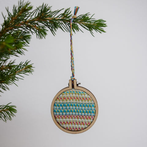 A Christmas bauble hangs from the sprig of a Christmas tree with a bright white background. The bauble is has a handwoven inner circular panel, surrounded in a ply embroidery hoop mount.
