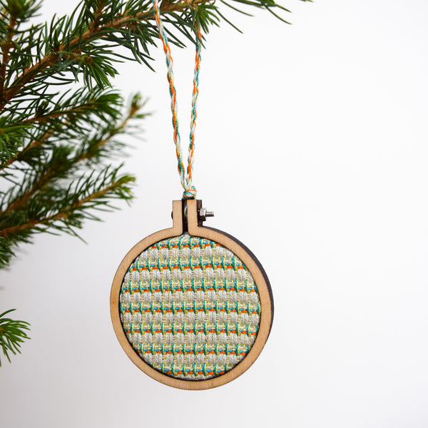 A Christmas bauble hangs from the sprig of a Christmas tree with a bright white background.  The bauble is has a handwoven inner circular panel, surrounded in a ply embroidery hoop mount.