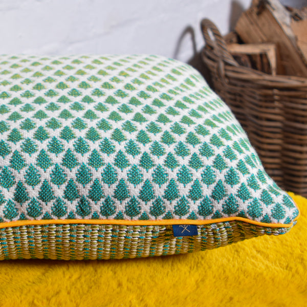 Picture of a handwoven cushion lying on a yellow fur mat.  The cushion has a design with dense green trees, becoming less dense and lighter as the pattern goes from bottom to top of the front panel.  A wicker basket with firewood can be seen in the background.