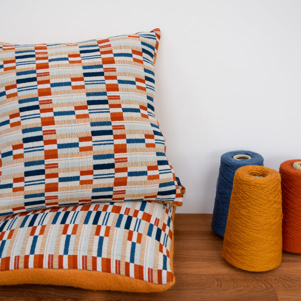 Photo showing showing two cushions sitting on top of each other with a mixture of horizontal rectangular blocks of orange, yellow and blues to create the design. There are also three cones of yarn showing the burnt orange, mustard and blue yarns which have been used to create the handwoven cloth.