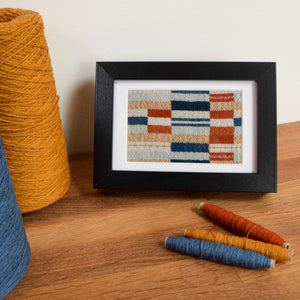 Handwoven art piece in black frame. Fabric is woven in blocks of varying colours of blues, yellows and burnt orange. In the foreground there are 3 bobbins with some blue, mustard and burnt orange yarns used within the woven design.