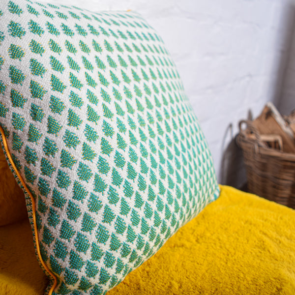 Picture of a handwoven cushion sitting on a yellow fur mat. The cushion has a design with dense green trees, becoming less dense and lighter as the pattern goes from bottom to top of the front panel. 