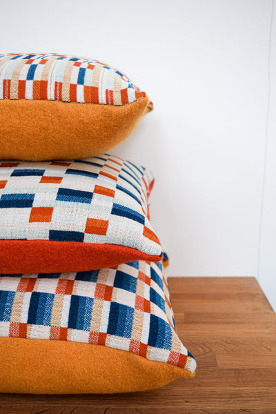 Three cushions piled in a stack on top of each other.  Each with a varying design of handwoven fabric with various shades of blue and orange block patterns.