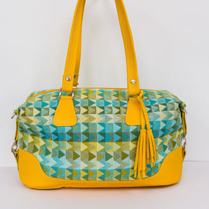Front facing image of a weekend bag.  Yellow leather base and trim with a handwoven triangular pattern fabric in varying shades of green, yellow and blue.