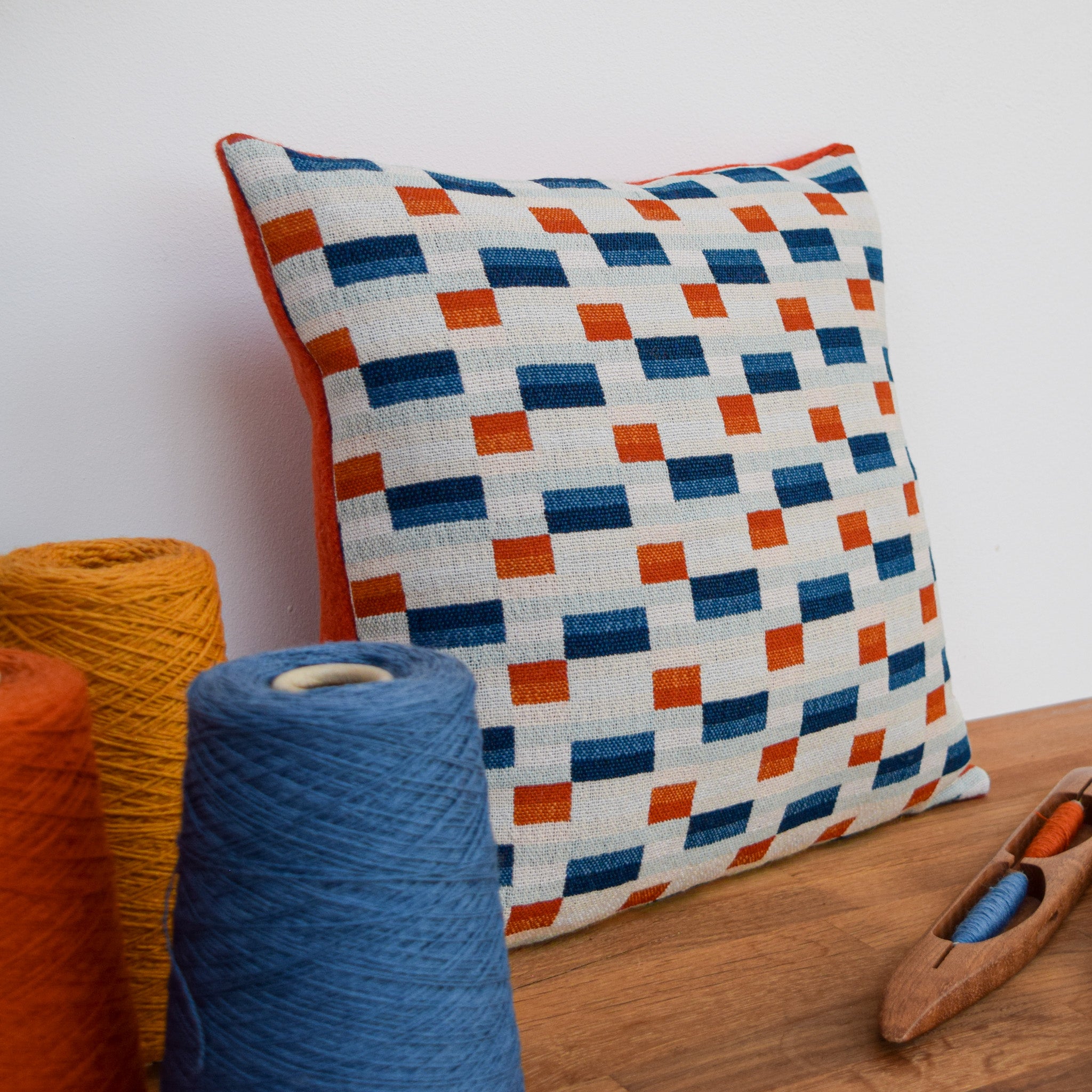 A handwoven cushion, designed with varying shades of blue, orange and buttercup rectangles sits in the centre.  It has a burnt orange boiled wool backing.  In the foreground sit 3 cones of weaving yarn showing the thread used within the design.  Alongside sits a wooden shuttle,  a tool used to weave the fabric.