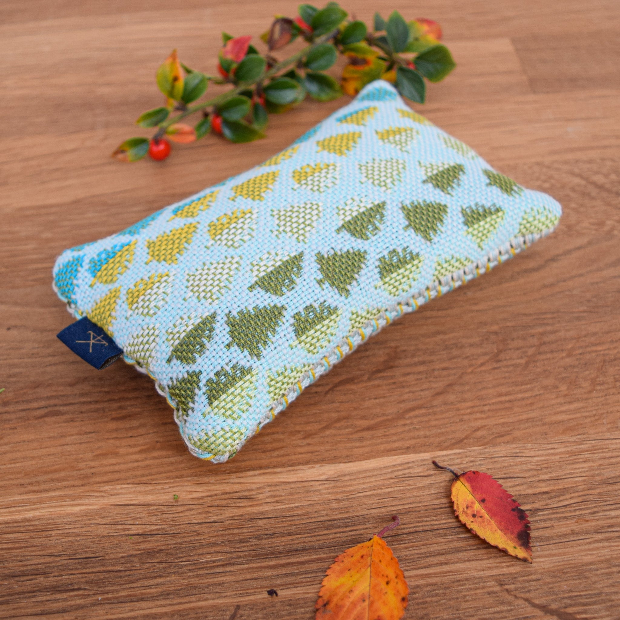 A small cedar pouch sitting on a wooden tabletop with fir tree handwoven design. Around the pouch are some autumnal leaves.