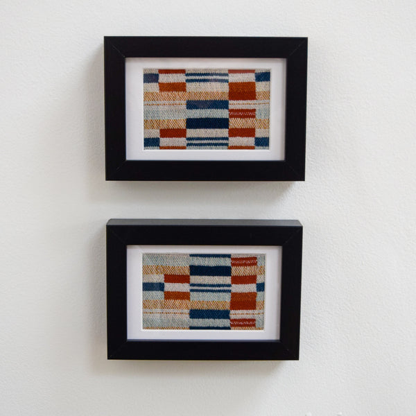 Two small black framed handwoven art pieces hung one above the other on a white wall.