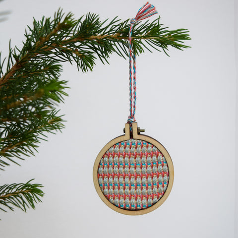 A Christmas bauble hangs from the sprig of a Christmas tree with a bright white background. The bauble is has a handwoven inner circular panel, surrounded in a ply embroidery hoop mount.