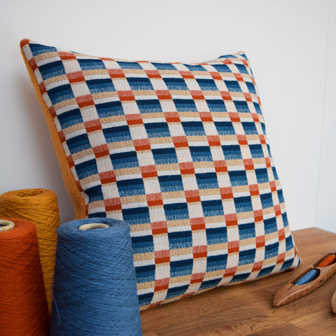 A handwoven cushion, designed with varying shades of blue, orange and buttercup rectangles sits in the centre. It has a burnt orange boiled wool backing. In the foreground sit 3 cones of weaving yarn showing the thread used within the design. Alongside sits a wooden shuttle, a tool used to weave the fabric.